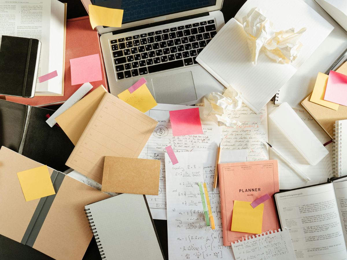 An overhead view of a cluttered desk with a laptop, scattered sticky notes, an open planner and notebooks